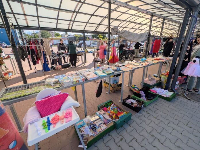 Stoisko z różnymi przedmiotami na miejskim targowisku z kupującymi w tle.