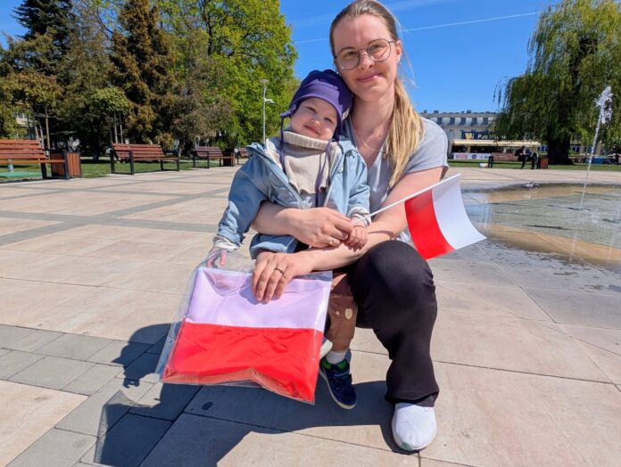 Kobieta trzymająca dziecko przed fontanną w parku, otoczona zielenią i wodnymi strumieniami.