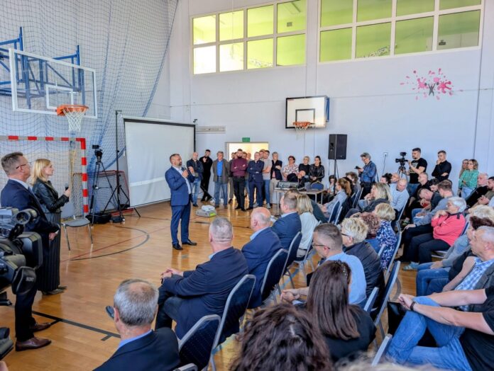 Grupa ludzi w sali sportowej obserwuje mężczyznę, który przemawia do mikrofonu