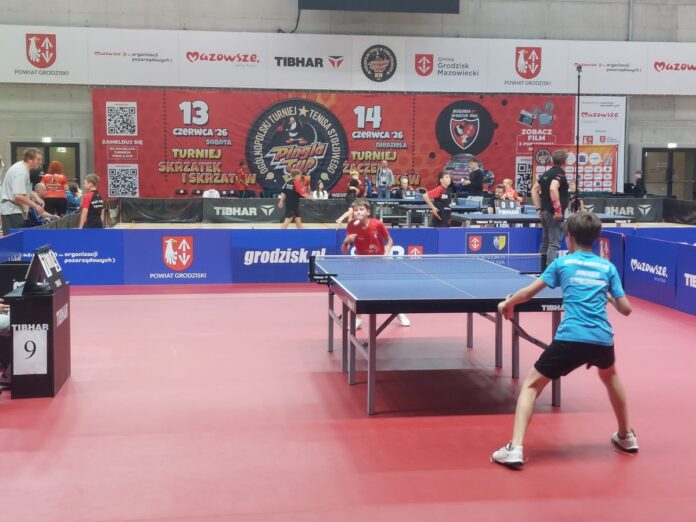 Dwaj młodzi mężczyźni grają w tenisa stołowego w dużej hali sportowej.