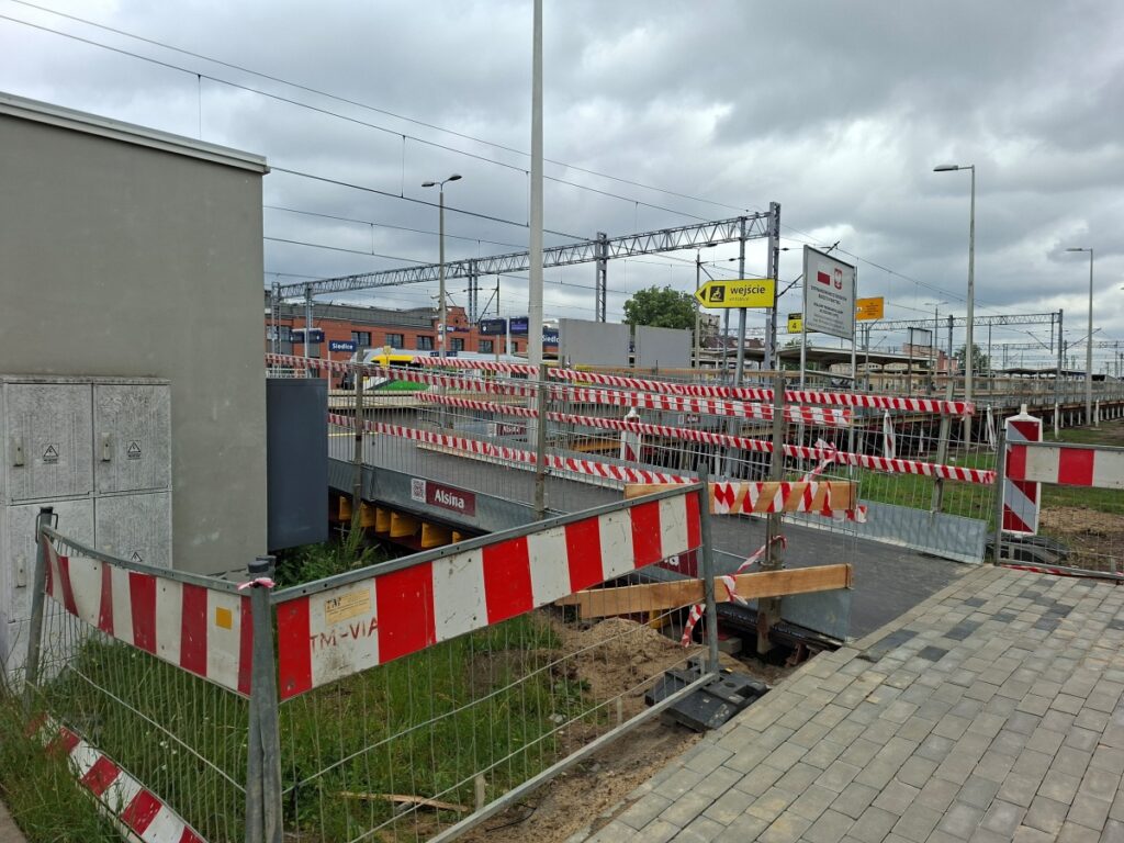Plac budowy z ogrodzeniem i drogą w tle, z widocznymi materiałami budowlanymi i sprzętem.