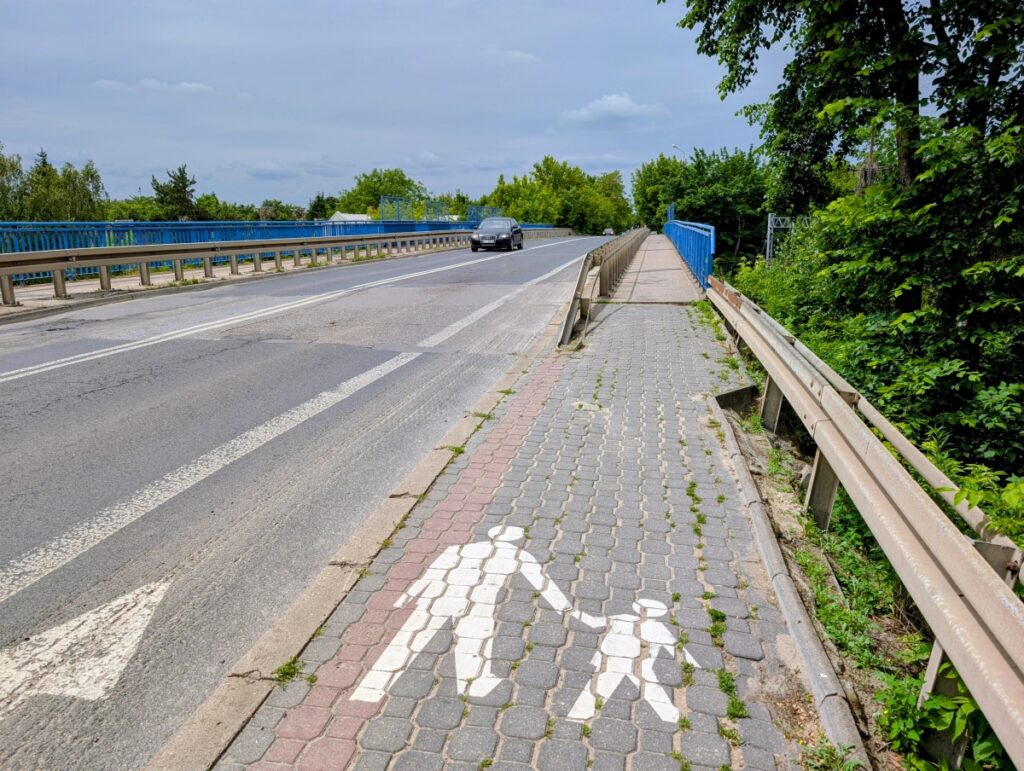 ulica i jadący po niej samochód, po obu stronach chodniki i niebieskie barierki