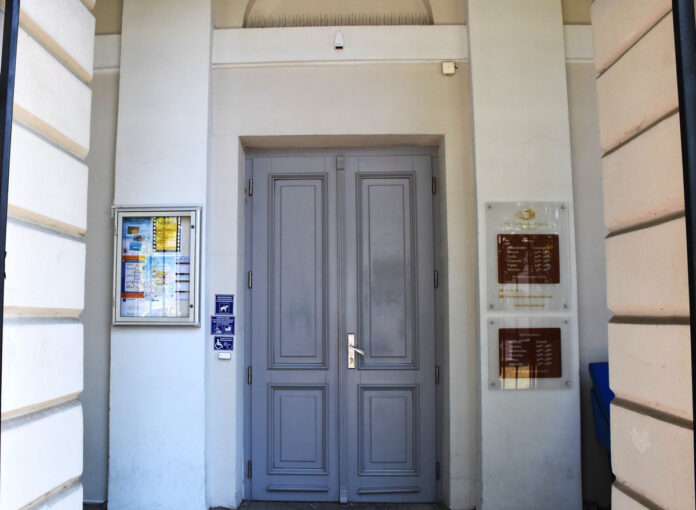 Szare dwuskrzydłowe drzwi wejściowe z tablicami informacyjnymi o godzinach otwarcia i ikonami dostępności