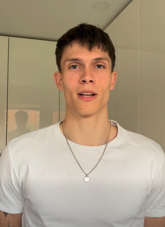 Młody mężczyzna z krótkimi brązowymi włosami, ubrany w biały t-shirt i srebrny łańcuszek