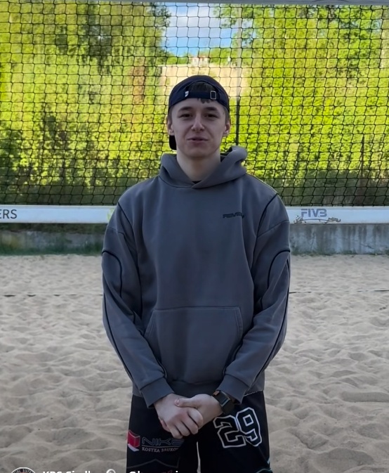 Młody mężczyzna w szarej bluzie z kapturem i czapce stoi na piaszczystym boisku przed siatką do siatkówki