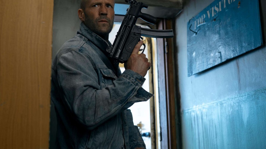Jason Statham w dżinsowej kurtce trzymający pistolet maszynowy w przejściu