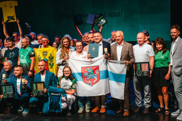 Grupa osób na scenie trzymająca nagrody, dyplomy oraz flagę Siedlec podczas uroczystej gali.