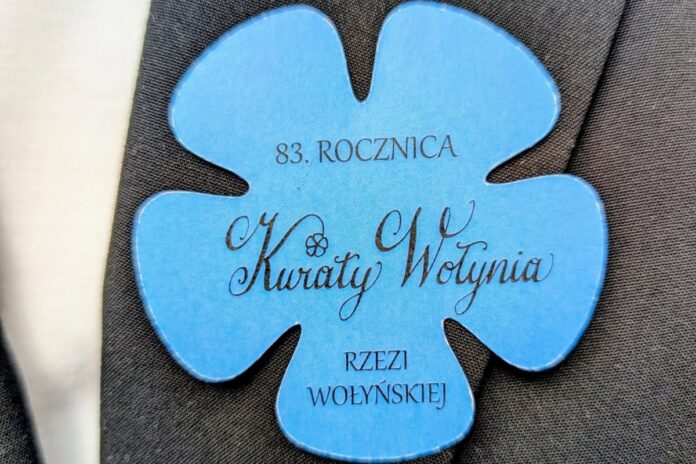 Niebieski kwiatek z napisem: 83. ROCZNICA Kwiaty Wołynia RZEZI WOŁYŃSKIEJ, przypięty do ciemnej marynarki