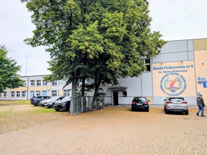 Dwukondygnacyjny budynek szkoły w pastelowych kolorach z drzewem pośrodku.