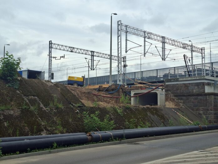 Tory kolejowe prowadzące w dal, otoczone zielonymi drzewami i krzewami w słoneczny dzień.