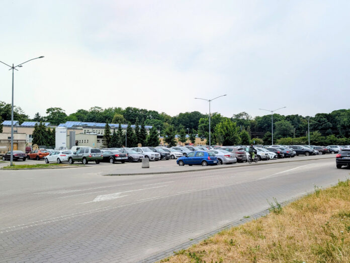Duży, zapełniony parking z wieloma samochodami przed budynkami i drzewami pod zachmurzonym niebem