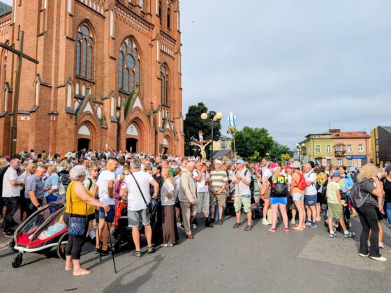 Tłum ludzi z krzyżem procesyjnym zgromadzony przed neogotyckim kościołem z czerwonej cegły