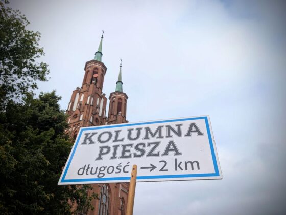 Tablica z napisem &bdquo;Kolumna piesza długość 2 km&rdquo; na tle wież ceglanego kościoła