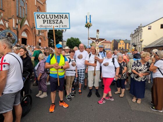 Pielgrzymi przed kościołem z tablicą &bdquo;KOLUMNA PIESZA długość -> 2 km&rdquo; oraz krzyżem procesyjnym