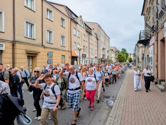 Grupa ludzi z plecakami idąca w kolumnie ulicą miasta, niekt&oacute;rzy machają do aparatu