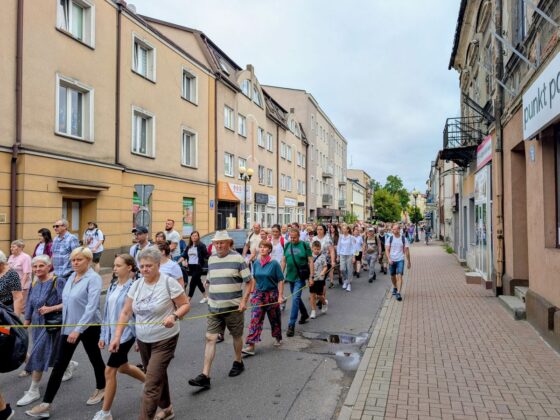 Grupa ludzi idąca ulicą miasta, trzymająca ż&oacute;łtą linę wyznaczającą kolumnę marszową