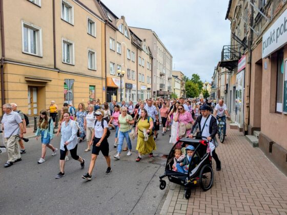 Tłum ludzi w r&oacute;żnym wieku idący środkiem miejskiej ulicy, w tym mężczyzna pchający podw&oacute;jny w&oacute;zek z dziećmi