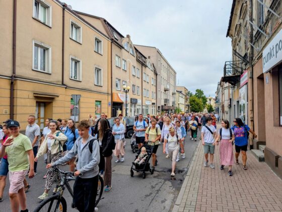 Tłum ludzi idący środkiem ulicy między kamienicami; niekt&oacute;rzy prowadzą rowery lub w&oacute;zki dziecięce
