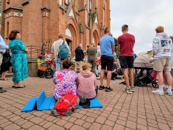 Grupa ludzi zgromadzona przed ceglanym kościołem; niekt&oacute;rzy siedzą na karimatach na bruku