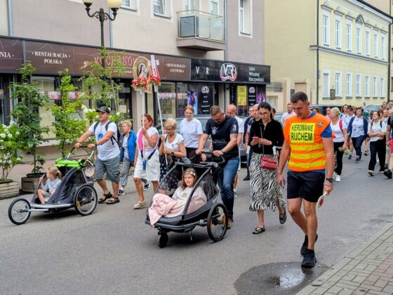 Grupa os&oacute;b w procesji na ulicy, w tym dzieci w w&oacute;zkach, kobieta z feretronem i mężczyzna kierujący ruchem
