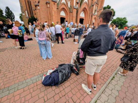 Tłum ludzi zgromadzony przed neogotyckim kościołem z czerwonej cegły, niekt&oacute;rzy siedzą na bruku