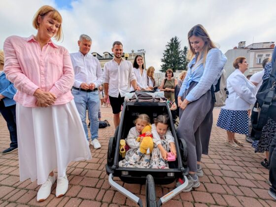 Grupa dorosłych uśmiecha się do dw&oacute;ch dziewczynek w w&oacute;zku; jedna z nich trzyma pluszowego Kubusia Puchatka