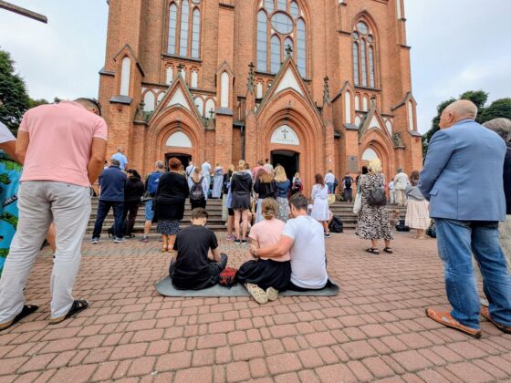 Tłum ludzi zgromadzony przed neogotyckim kościołem z czerwonej cegły; para siedzi na macie na pierwszym planie