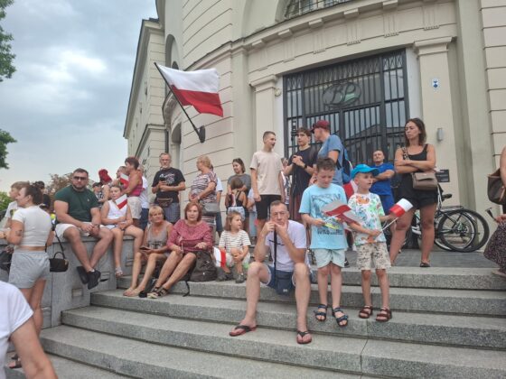 Grupa ludzi z polskimi flagami zgromadzona na schodach budynku