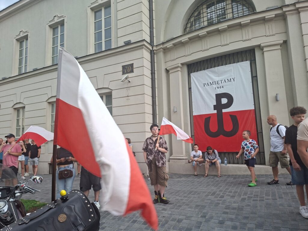 Ludzie z polskimi flagami przed banerem z symbolem Polski Walczącej i napisem Pamiętamy 1944