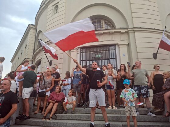 Grupa ludzi z polskimi flagami na schodach budynku; w centrum mężczyzna machający dużą flagą
