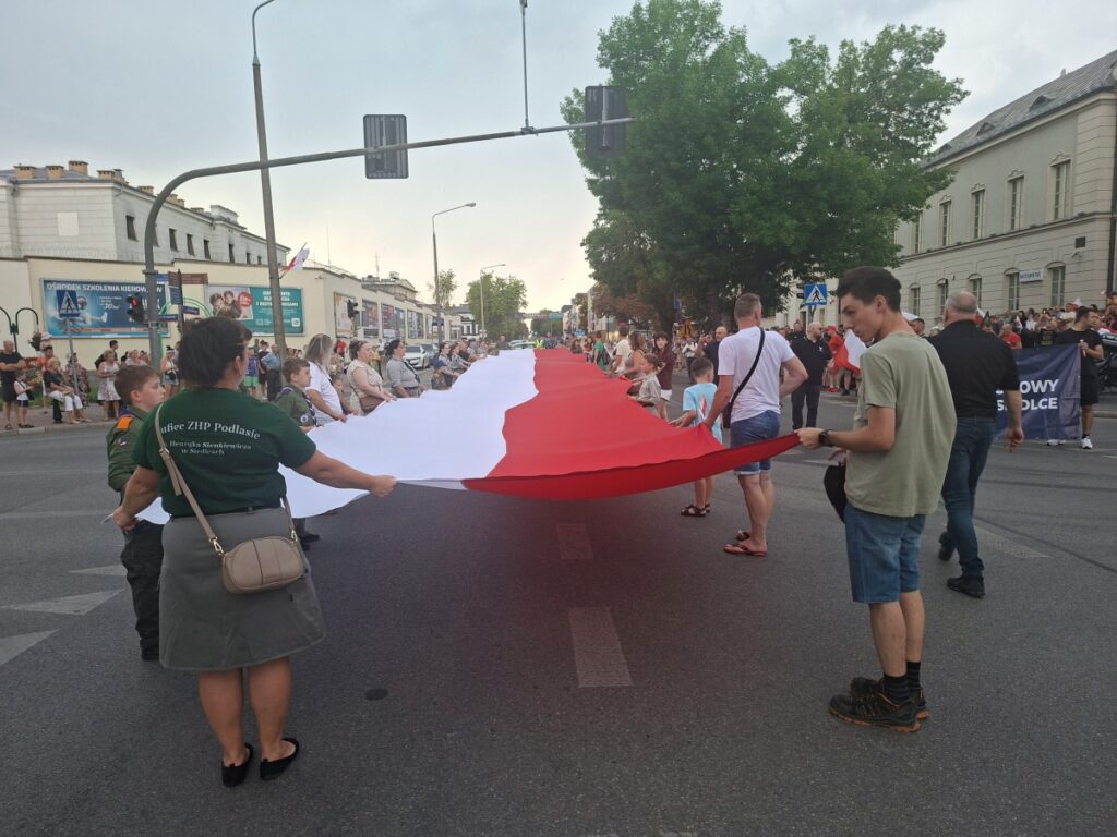 Grupa ludzi, w tym harcerze, trzymająca rozciągniętą, długą polską flagę na środku ulicy