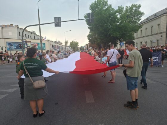 Grupa ludzi, w tym harcerze, trzymająca rozciągniętą, długą polską flagę na środku ulicy