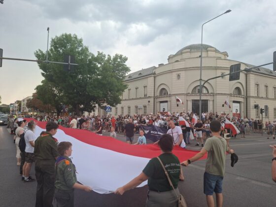 Tłum ludzi, w tym harcerze, trzymający wielką polską flagę na ulicy podczas uroczystości patriotycznych
