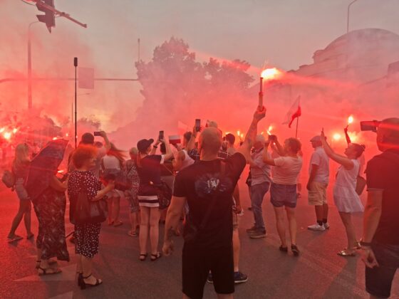Tłum ludzi na ulicy z zapalonymi czerwonymi flarami i polskimi flagami w gęstym, czerwonym dymie
