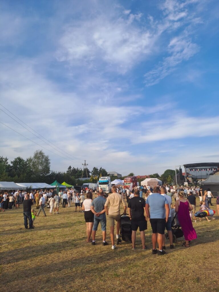 Tłum ludzi na festynie plenerowym z namiotami, ciężarówkami i sceną w tle pod błękitnym niebem