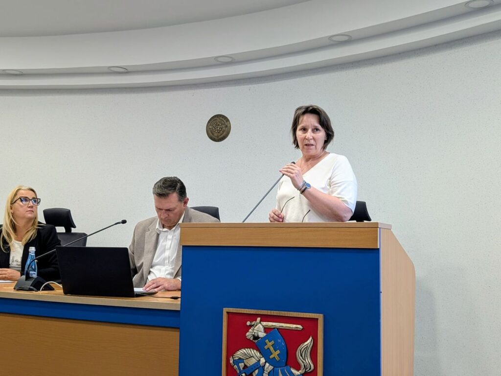 Kobieta przemawiająca przy mównicy z mikrofonem podczas oficjalnego wystąpienia. Obok przy stole siedzą dwie osoby