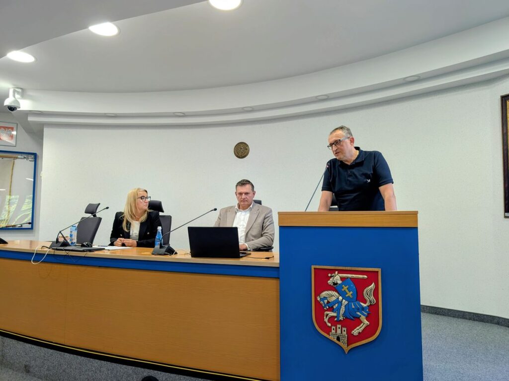 Mężczyzna przemawia przy mównicy z herbem Siedlec, obok dwie osoby siedzące przy stole prezydialnym
