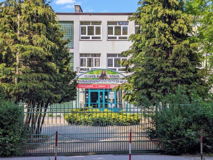 Wejście do Zespołu Szkół Ponadpodstawowych nr 4 za metalowym ogrodzeniem w otoczeniu wysokich drzew