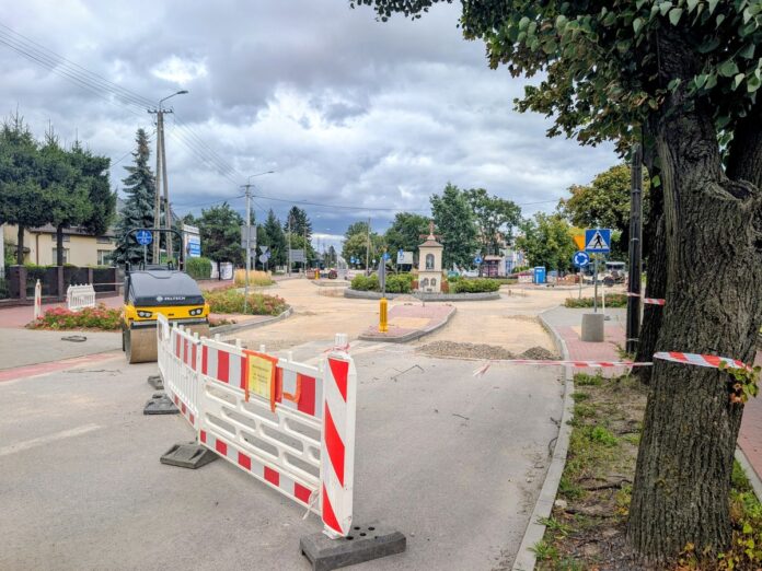 Plac budowy drogi z biało-czerwonymi barierami, walcem drogowym i kapliczką na rondzie w tle