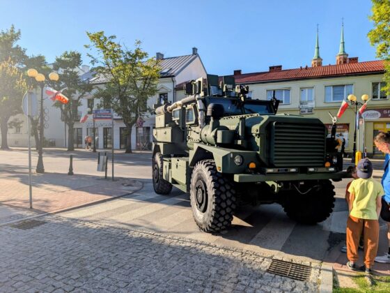 Opancerzony pojazd wojskowy zaparkowany na miejskiej ulicy, obok kt&oacute;rego stoją przechodnie