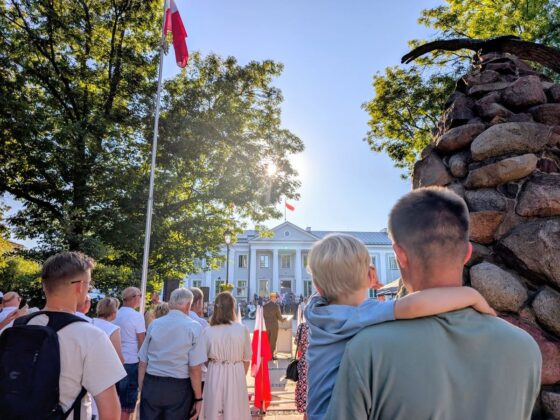 Uroczystość patriotyczna przed klasycystycznym budynkiem; zgromadzeni ludzie, polskie flagi i żołnierz przy m&oacute;wnicy