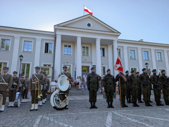 Orkiestra wojskowa i żołnierze w mundurach polowych przed klasycystycznym budynkiem z polską flagą