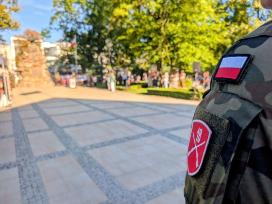 Ramię żołnierza z polską flagą i naszywką jednostki na tle uroczystości patriotycznej