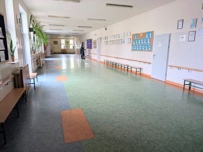 Jasny, szeroki korytarz szkolny z zieloną podłogą, ławkami i dekoracjami na ścianach