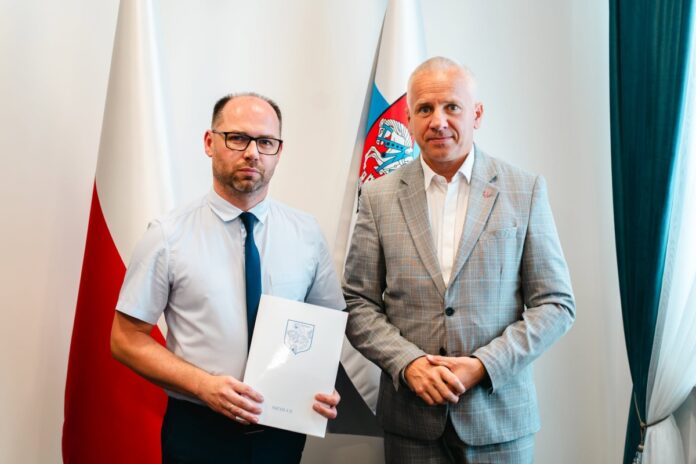 Dwóch mężczyzn w strojach wizytowych przed flagami Polski i Siedlec; jeden trzyma teczkę z napisem Siedlce