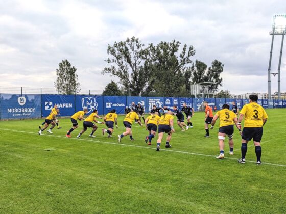 Mecz rugby na trawiastym boisku, w kt&oacute;rym zawodnicy w ż&oacute;łtych strojach biegną w stronę drużyny w czarnych koszulkach