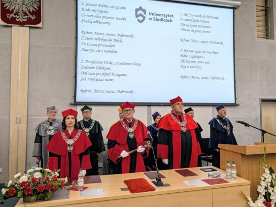 Władze Uniwersytetu w Siedlcach w togach akademickich na scenie podczas uroczystości. W tle ekran z hymnem Polski