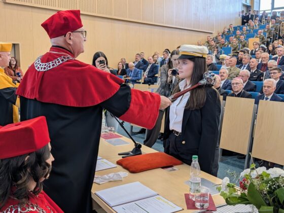 Rektor w czerwonej todze pasuje studentkę berłem podczas uroczystości akademickiej w auli