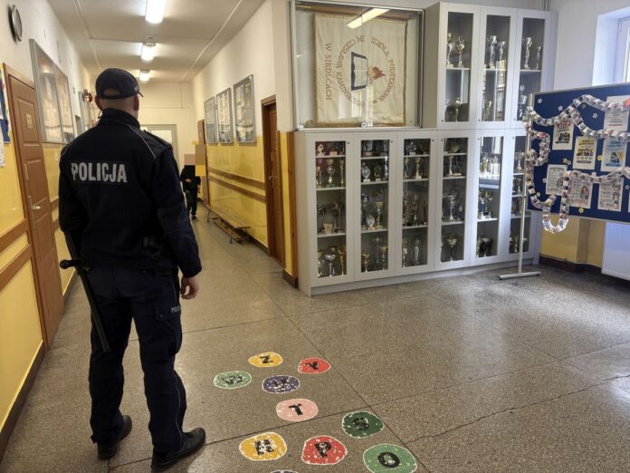 Policjant stojący na korytarzu szkoły podstawowej obok gablot z pucharami